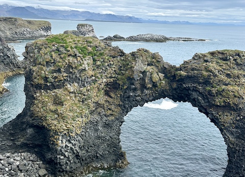 A natural rock arch formation along a coastal landscape, surrounded by water and scenic mountains in the background.