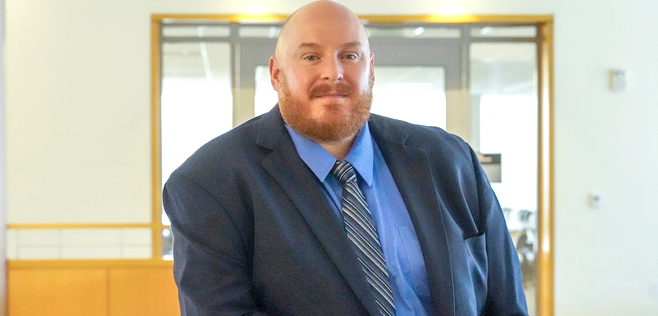 John Lauck, smiling, wearing a blue dress shirt, striped tie, and dark suit jacket in an office hallway.