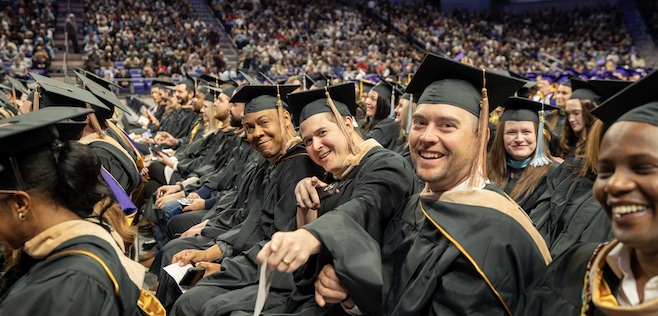 A group of smiling JMU MBA graduates in caps and gowns celebrating during a graduation ceremony.