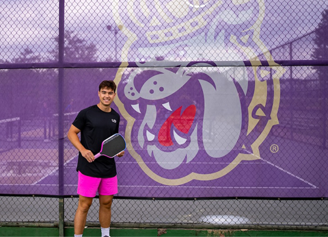 Ryan Morneau holding a pickleball paddle in front of a purple fence banner with the Duke bulldog logo on an outdoor court.