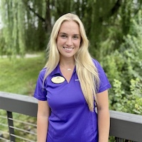 A smiling young woman in a purple polo shirt stands outdoors with greenery in the background.