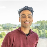 A smiling young man in a purple polo shirt stands on a balcony, surrounded by greenery.