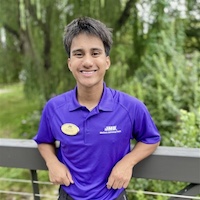 A smiling young man in a purple polo shirt stands on a balcony, surrounded by greenery.