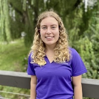 A smiling young woman with curly hair wearing a purple shirt stands outdoors against a backdrop of greenery.
