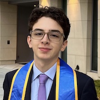 Andres Parra wearing glasses, a suit, and a graduation sash poses confidently outdoors.