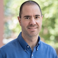 Professional headshot of a man with short hair, wearing a blue button-up shirt and smiling at the camera.