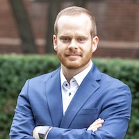 Professional headshot of a man in a blue suit, standing outdoors with arms crossed, exuding confidence.