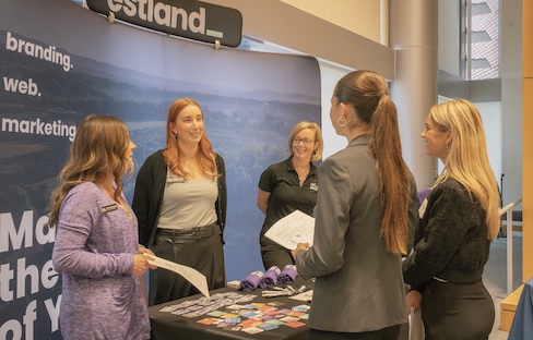 Four women talk at an exhibitor booth with brochures and promotional items on a table in a conference hall.