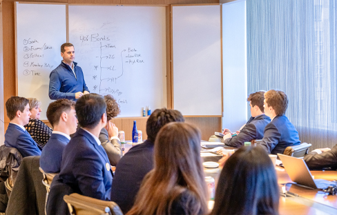 A group of students sit at a long conference table while a man presents in the front of the room.