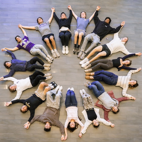 A group of people lying on the floor in a circular formation, holding hands and smiling.