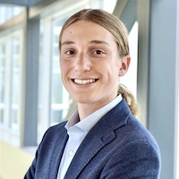 Smiling young man in a blazer, standing in a sunlit interior space.