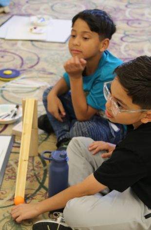 two kids sitting on the floor, rolling a ball down a wooden ramp