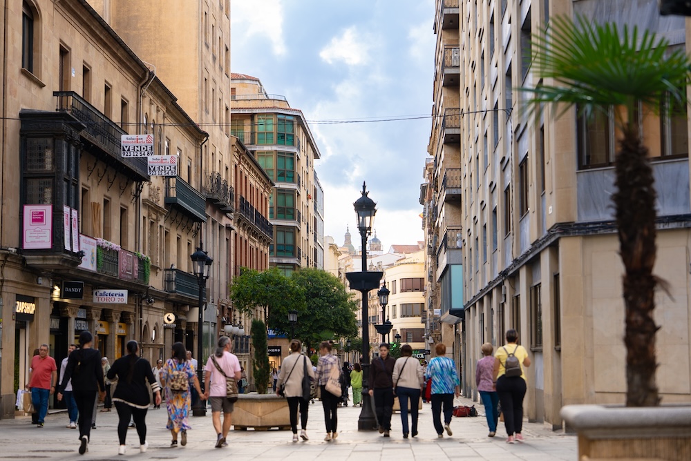 A bustling urban street scene with pedestrians walking among shops and tall buildings, framed by greenery and lampposts.