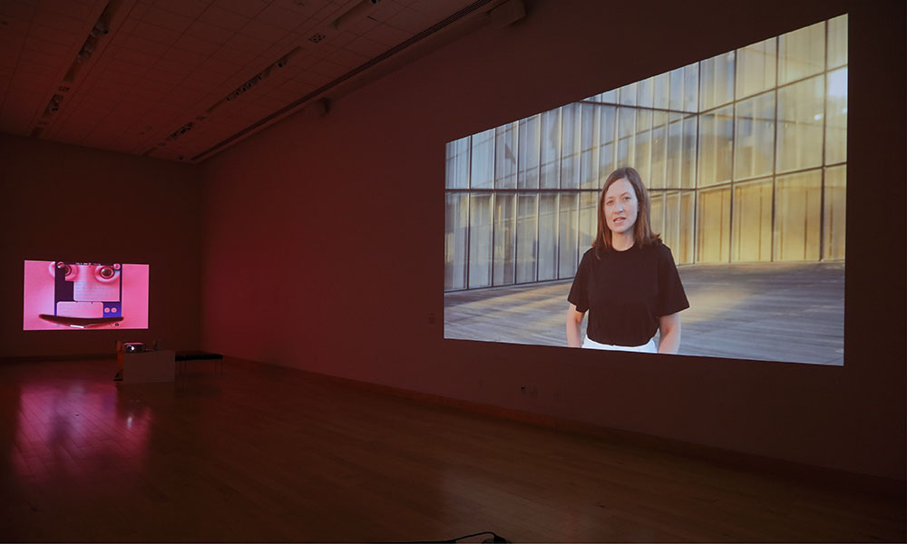 A still of a woman speaking in front of a glass building is projected onto a wall on the right and on the left is a pink projection of an artwork. The work features human eyes and text messages.