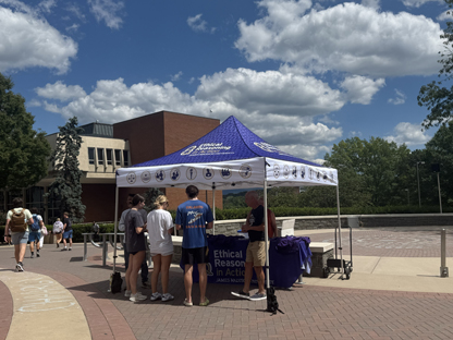 A group of people gathering at a informational booth under a tent on a sunny day, with a sign promoting ethical reasoning and engagement.