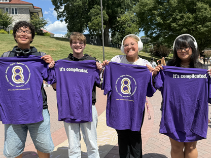 A group of four students wearing purple t-shirts that read "It's complicated." standing outdoors on a college campus.