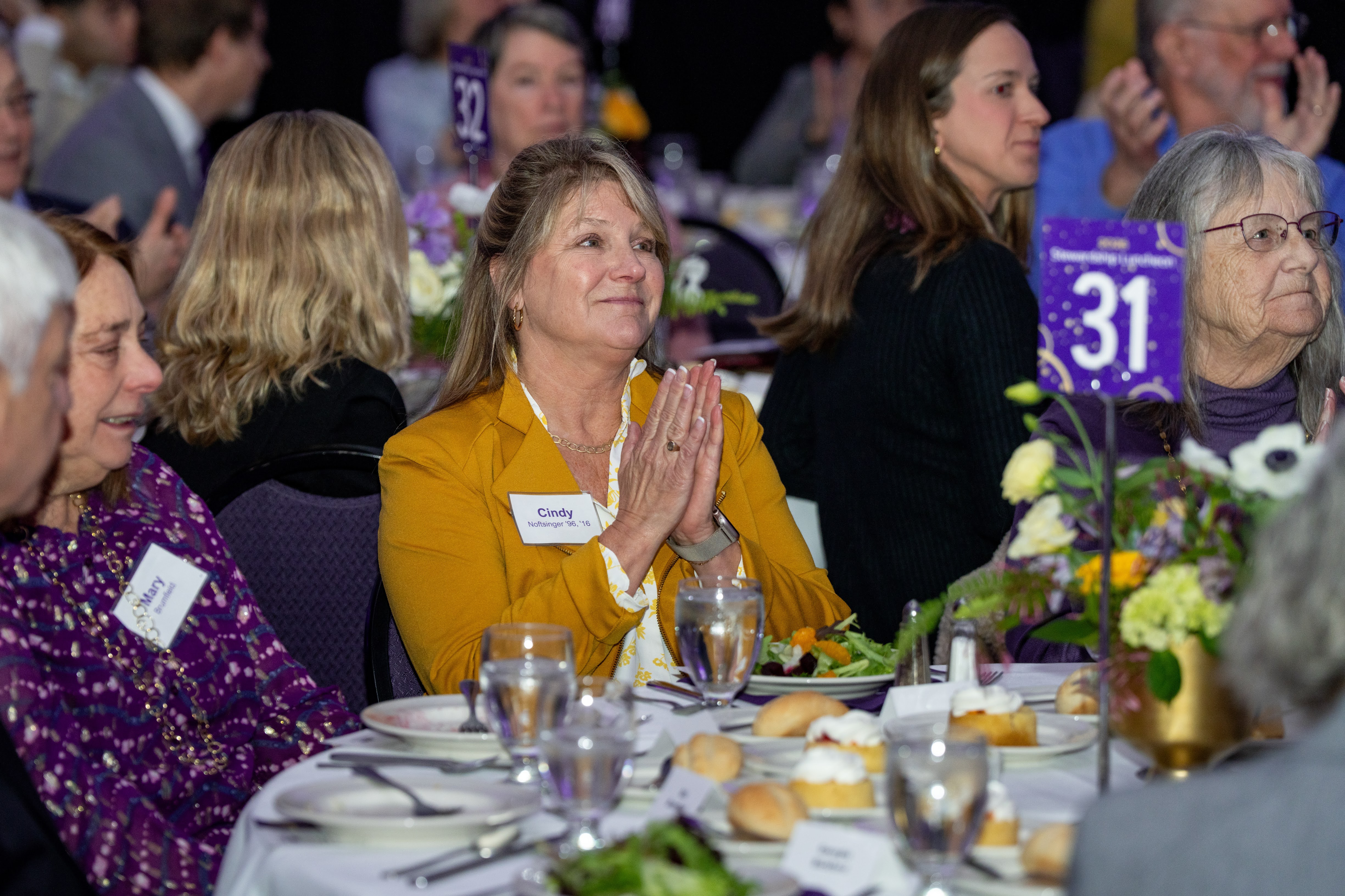 Audience members applaud at a banquet table with floral centerpieces and numbered table signs.