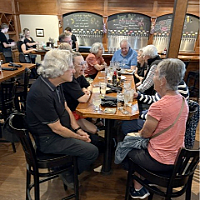 Group of older adults gathered around a wooden table in a restaurant, sharing food and drinks with a bar and chalkboard menu in the background.