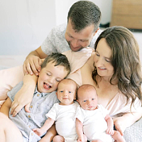 Smiling parents cuddle their three young children—an older toddler and two babies—while sitting together on a bed.