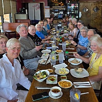 Large group of older adults seated around a long table at a restaurant, smiling with plates of food and drinks in front of them.