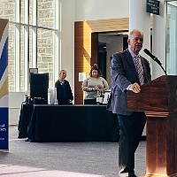 Older man in a suit speaks at a wooden podium in a bright event space, with attendees and a registration table in the background.