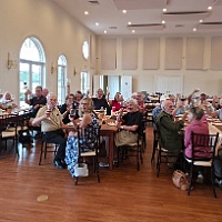 Seniors seated at tables in a bright dining hall with large arched windows, socializing during a group event.