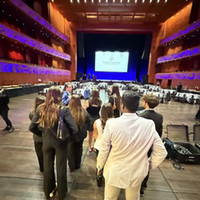 A group of people in formal attire walking towards a stage in a large auditorium setting, with empty tables set up in the foreground.