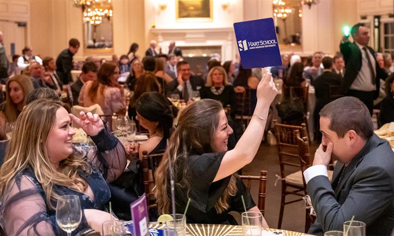 A group of people are seated around a large, curved conference table during the Le Gourmet live auction event. The setting is a bright, modern room with tall floor-to-ceiling windows that reveal trees with autumn foliage outside. Several attendees are visible from behind in the foreground, while three individuals are clearly seen across the table, engaged in conversation. The table is organized with nameplates, papers, and water bottles, and the chairs are sleek, high-backed office chairs. The atmosphere appears professional and collaborative, reflecting a formal auction
