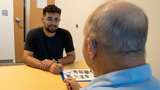 Young man with glasses talks with an older man across a table while reviewing a clipboard of photos.