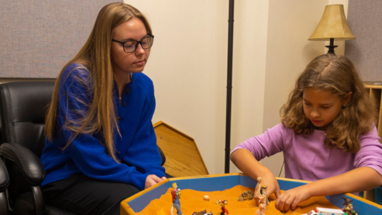 Woman sits beside a young girl as she plays with figurines in a sandbox-style play tray in a therapy or counseling room.