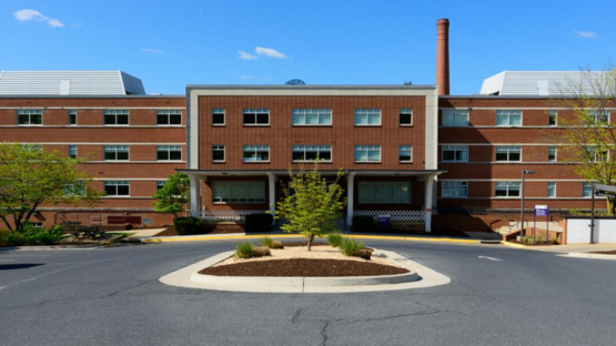 Brick institutional building with a central entrance and a tall smokestack, viewed from a circular driveway under a clear blue sky.