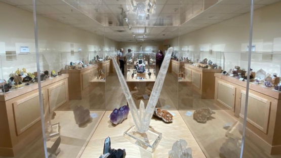 Glass display case in a museum mineral gallery, featuring large clear crystal points and colorful mineral specimens on shelves in the background.