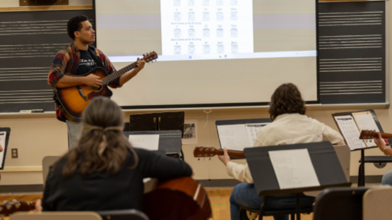 Guitar instructor demonstrating chords at the front of a classroom while students practice with guitars and music stands.