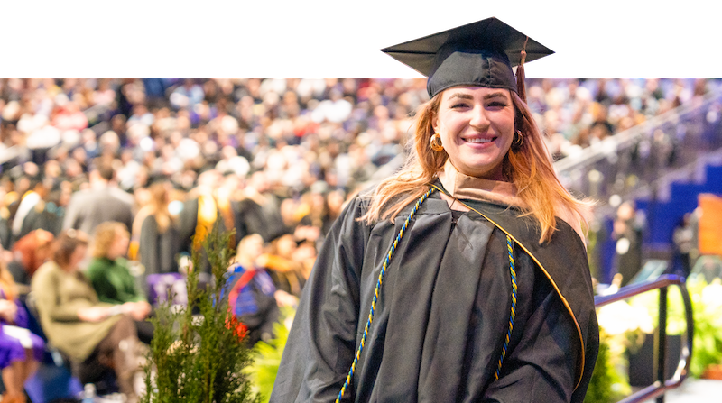 A smiling graduate in a black cap and gown celebrates at a commencement ceremony, with fellow graduates seated in the background.