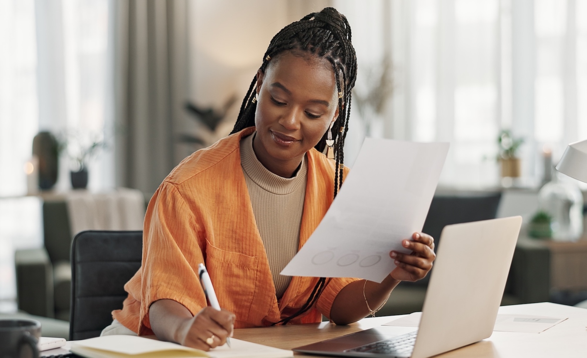 A woman in an orange cardigan sits at a desk, reviewing documents and taking notes while using a laptop.