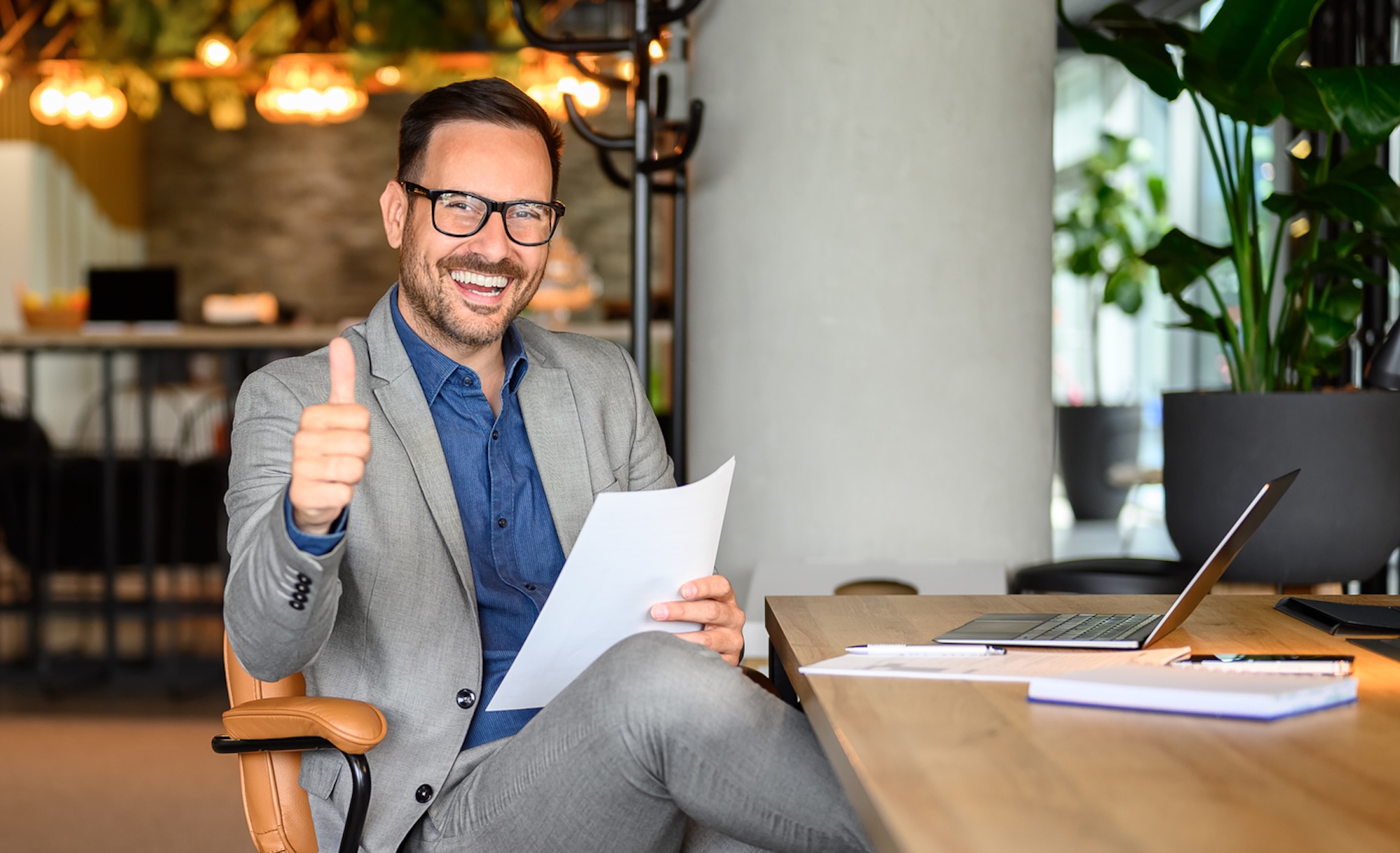 Smiling professional man in a suit gives a thumbs-up while sitting at a desk with a laptop and documents.