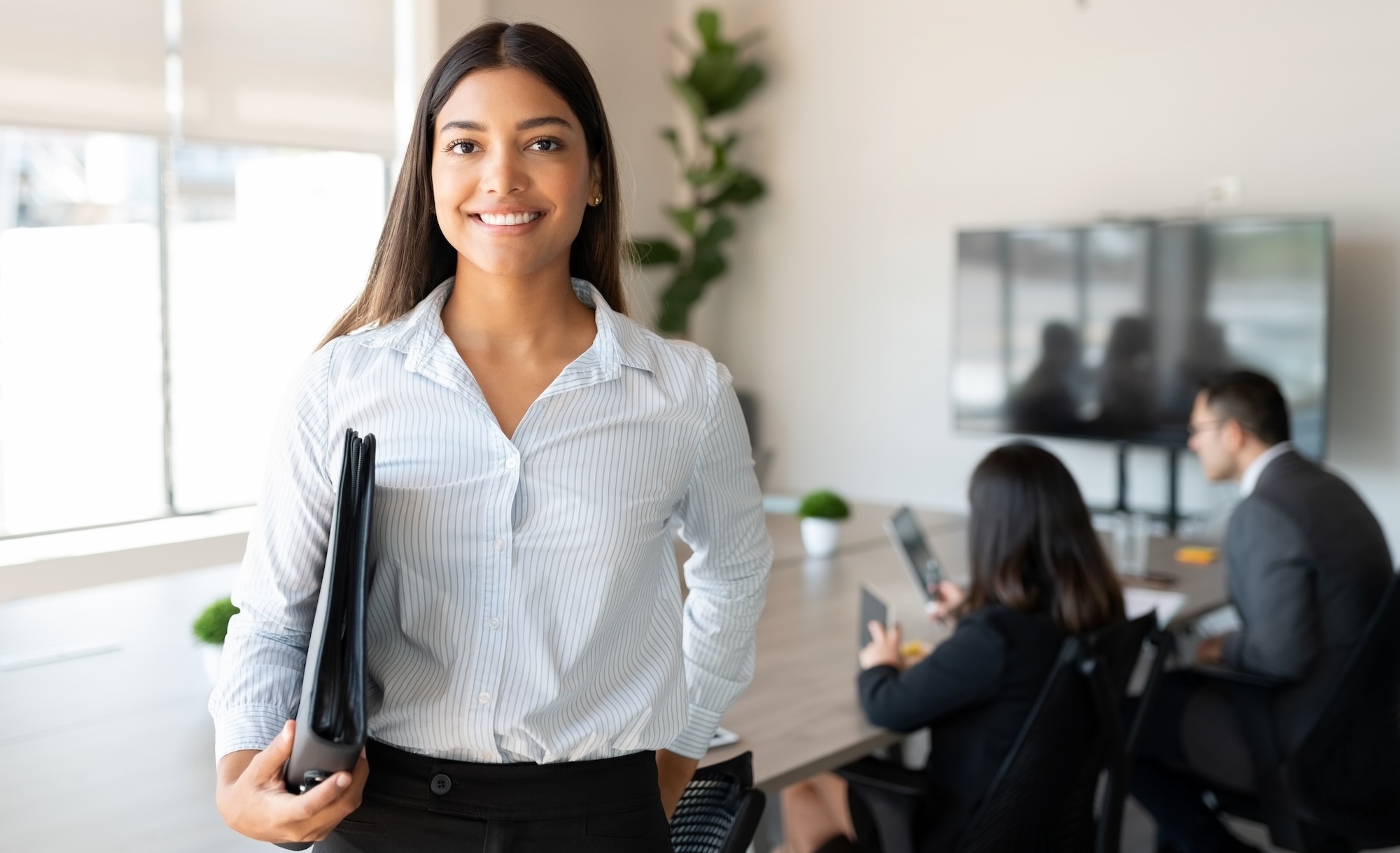 A confident businesswoman stands in a modern office setting, holding a clipboard, with colleagues engaged in a meeting in the background.