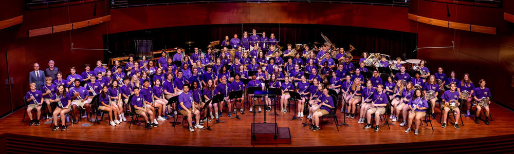 Large school concert band in purple shirts posing on an auditorium stage with their instruments.