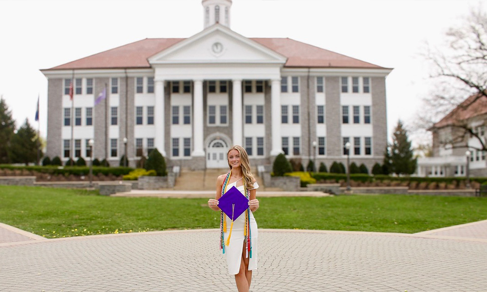 Abby Killmon in a white dress holding a purple cap and surrounded by graduation cords stands in front of Wilson Hall.
