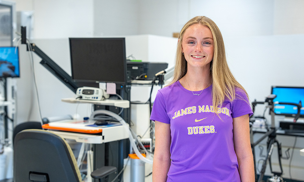 Abby Killmon wears a purple James Madison University t-shirt stands in a modern clinical lab setting filled with medical equipment.