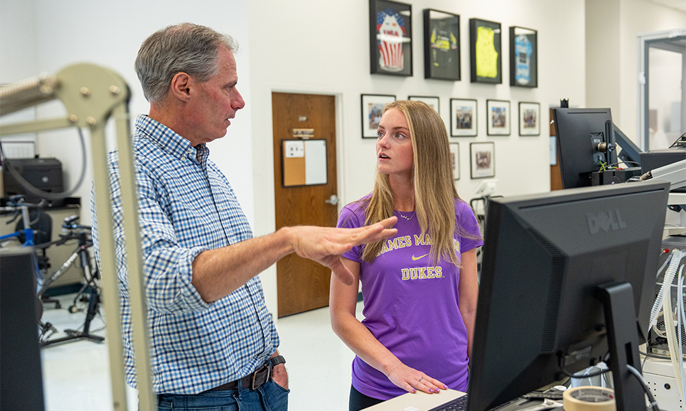Dr. Chris Womack discusses research with Abby Killmon in a laboratory setting, featuring various equipment and framed photographs in the background.