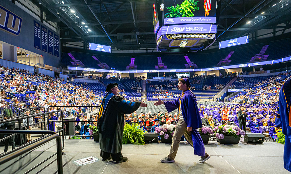 A graduate in cap and gown walks across stage to shake hands with a faculty member.