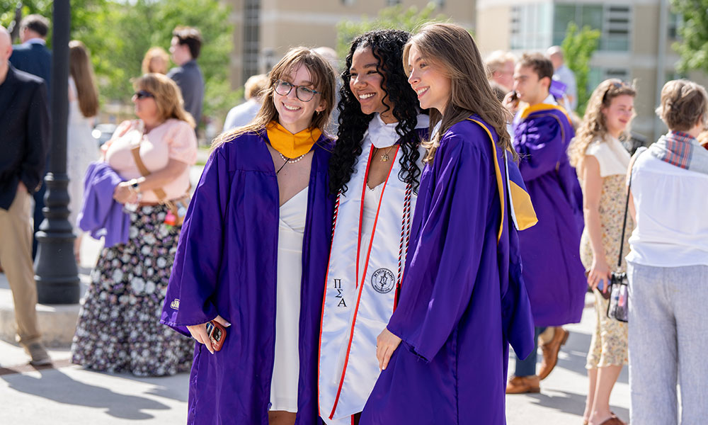 Three graduates in purple gowns pose together outdoors, smiling and celebrating their achievement.