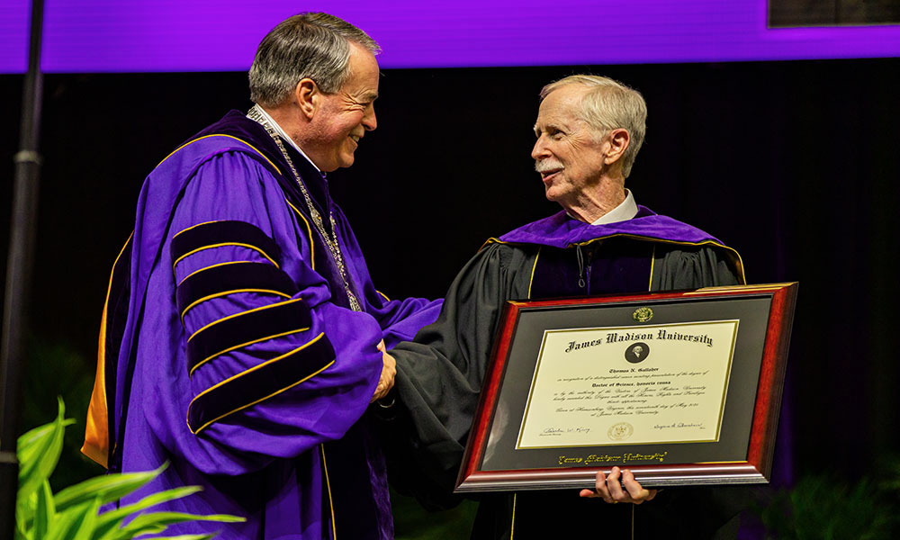 A man in academic regalia presents a diploma to another man in a graduation gown and hood during a ceremony at James Madison University.