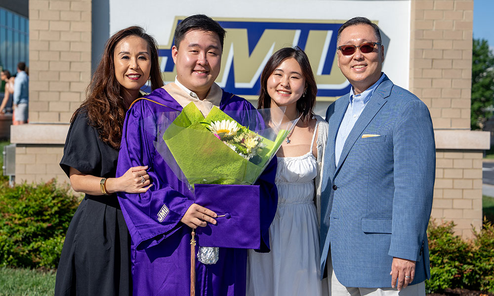 A proud graduate in a purple gown holds a bouquet while posing with his family outside a university building.