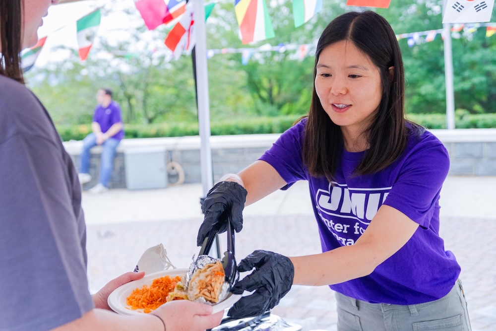 A woman in a purple JMU t-shirt serves food to a guest at an outdoor event decorated with international flags.
