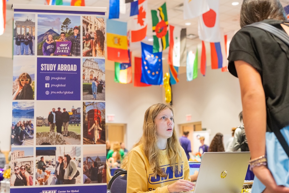 A student representative at a study abroad fair engages with attendees at a booth featuring a banner showcasing various international experiences.