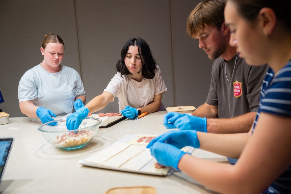 A group of four individuals engaged in a cooking activity, preparing food on a countertop while wearing blue gloves.