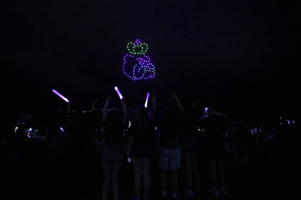 Crowd at a nighttime waving purple glow sticks while a drone light display forms a Duke dog in the sky.