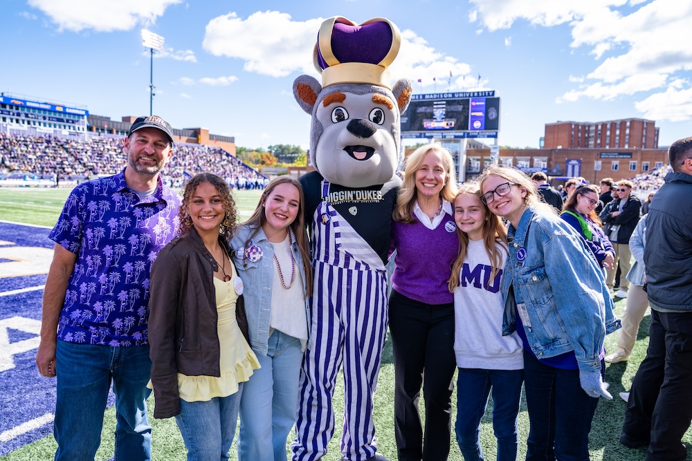 Group of people pose with Duke Dog wearing a purple-and-white striped overalls on a football field, with stadium stands in the background.